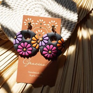May include: Black, round earrings with colorful flower designs in pink, orange, and purple. Each earring has a gold-colored stud and a small gold accent in the center of each flower. The earrings are displayed on a brown card with the word "Gracias!" and "Handmade by Karla Vega".