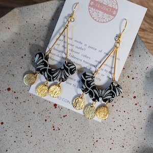 Puede incluir: Pendientes de cerámica negra y blanca con un diseño geométrico y detalles dorados. Los pendientes cuelgan de un alambre dorado.