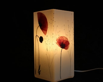 May include: A rectangular table lamp with a translucent white shade featuring pressed red poppy flowers and thin dark stems. The lamp emits a warm internal glow, highlighting the delicate botanical details against the light surface.