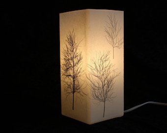May include: A rectangular table lamp with a translucent white shade featuring a dark brown botanical print of three distinct tree-like silhouettes. The lamp emits a warm, soft glow from within, highlighting the intricate branch details.
