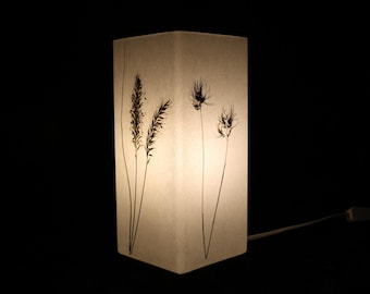 May include: A rectangular table lamp with a translucent white shade featuring a black botanical print of tall grasses and seed heads. The lamp emits a soft glow and includes a white power cord with an inline switch extending from the base.