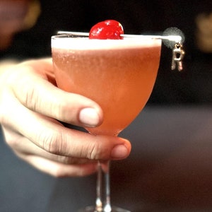 May include: A pink cocktail with a maraschino cherry garnish in a stemmed glass. The glass is being held by a hand.