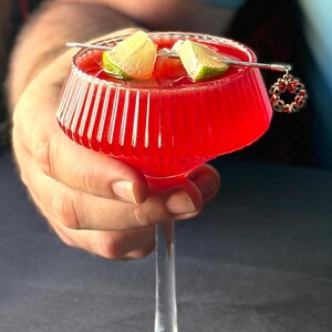 May include: A vibrant red cocktail in a stemmed glass, garnished with lime wedges. A decorative charm hangs from the cocktail pick. The glass has a ribbed design and is held by a person.