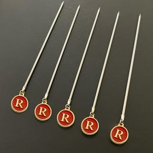 May include: Set of five silver cocktail stirrers with red and gold round charms. Each charm has a gold letter 'R' on a red background.
