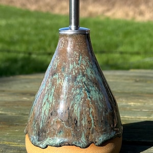 May include: A ceramic oil bottle with a stainless steel spout. The bottle has a brown and teal glaze over a tan base. The bottle is pear-shaped and has a shiny finish. The spout has a small lever to control the flow of oil.