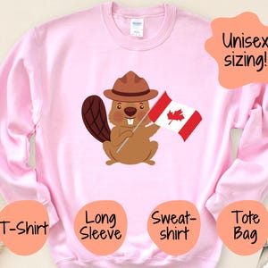 May include: Pink long-sleeve sweatshirt with a cartoon beaver wearing a ranger hat and holding a Canadian flag. The text "Unisex sizing!" is in a speech bubble above the beaver. The sweatshirt is available in T-shirt, long sleeve, sweatshirt, and tote bag options.