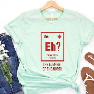 May include: A light green t-shirt with a red graphic of a periodic table element. The element is labeled "Eh?" and "Canadium [18.670]" with a Canadian flag icon. The text below the element reads "The Element of the North".