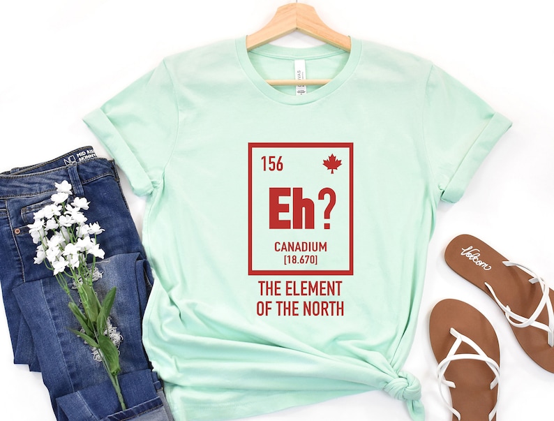 May include: A light green t-shirt with a red graphic of a periodic table element. The element is labeled "Eh?" and "Canadium [18.670]" with a Canadian flag icon. The text below the element reads "The Element of the North".