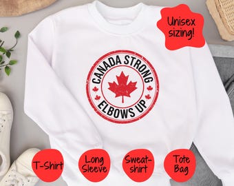 May include: White sweatshirt with a red circle graphic that says "Canada Strong Elbows Up" with a red maple leaf inside the circle. The sweatshirt is available in a variety of styles, including a t-shirt, long sleeve, sweatshirt, and tote bag.