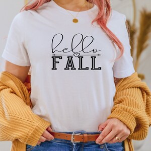 以下が含まれることがあります： 黒い筆記体で「hello fall」と書かれた白いTシャツ。矢印とハートが「fall」の上にあります。