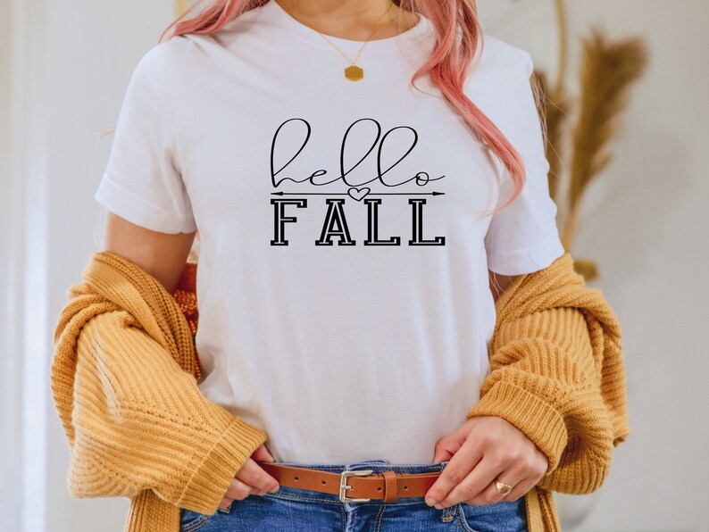 以下が含まれることがあります： 黒い筆記体で「hello fall」と書かれた白いTシャツ。矢印とハートが「fall」の上にあります。