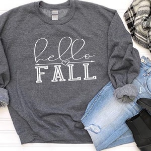 以下が含まれることがあります： 白い文字で「hello fall」と書かれたグレーのスウェットシャツ。小さなハートと矢印のグラフィックが描かれている。