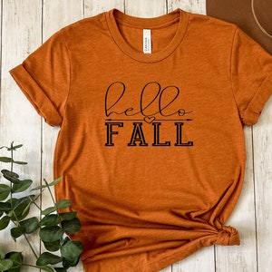 以下が含まれることがあります： さび色のTシャツに、黒い文字で「hello fall」と書いてあり、小さなハートと矢印のグラフィックが描かれています。