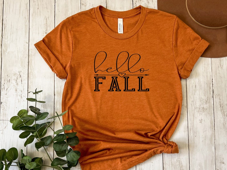 以下が含まれることがあります： さび色のTシャツに、黒い文字で「hello fall」と書いてあり、小さなハートと矢印のグラフィックが描かれています。
