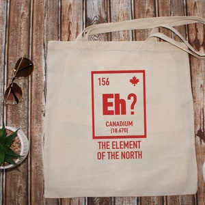 May include: A white canvas tote bag with a red graphic of a periodic table element. The element is labeled "Eh?" with the text "Canadium [18.670] The Element of the North".