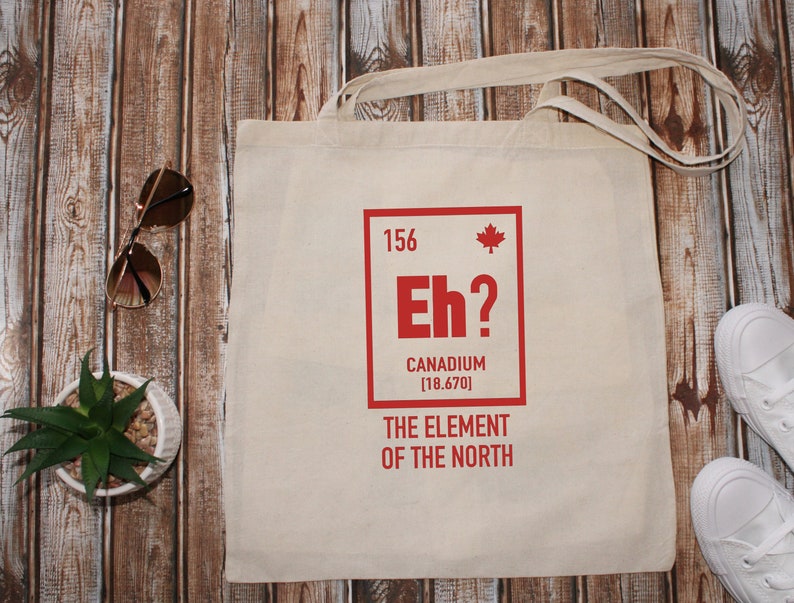 May include: A white canvas tote bag with a red graphic of a periodic table element. The element is labeled "Eh?" with the text "Canadium [18.670] The Element of the North".