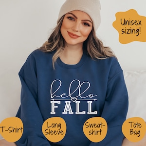 May include: A blue long-sleeved sweatshirt with the text "hello FALL" in white lettering. The sweatshirt has a small heart design above the word "FALL". The text is in a handwritten font. The sweatshirt is unisex sizing.