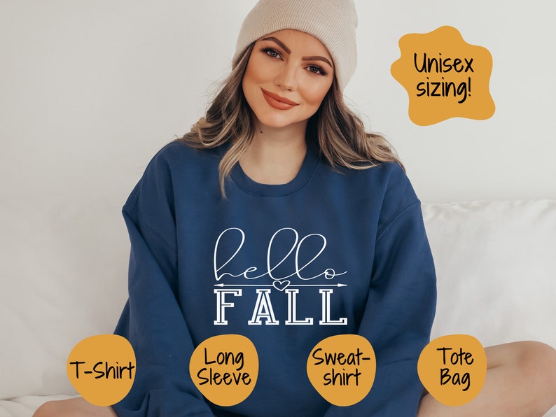 以下が含まれることがあります： 白い文字で「hello FALL」と書かれた青い長袖スウェットシャツ。スウェットシャツは「FALL」の文字の上に小さなハートのデザインがあります。テキストは手書きのフォントです。スウェットシャツはユニセックスサイズです。