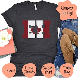 May include: A dark gray t-shirt with a white and red graphic of the Canadian flag. The graphic is stylized to look like the letters "EH". The t-shirt is unisex sizing. The image also shows a tote bag, long-sleeve shirt, and sweatshirt.