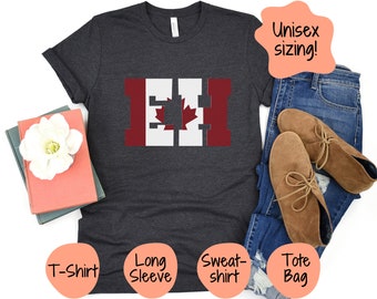 May include: A dark gray t-shirt with a white and red graphic of the Canadian flag. The graphic is stylized to look like the letters "EH". The t-shirt is unisex sizing. The image also shows a tote bag, long-sleeve shirt, and sweatshirt.