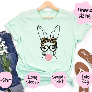 May include: A light green t-shirt featuring a black and white illustration of a bunny wearing leopard print sunglasses and a headband. The bunny is blowing a pink bubble. The text "Unisex sizing!" is in a pink speech bubble. The t-shirt is available in a variety of styles, including long sleeve and sweatshirt.