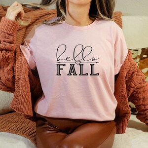 以下が含まれることがあります： 黒い文字で「hello fall」と書かれたピンクのTシャツ。文字の上と下に小さなハートと矢印があります。