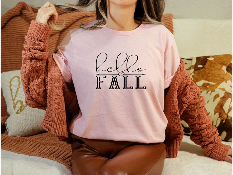 以下が含まれることがあります： 黒い文字で「hello fall」と書かれたピンクのTシャツ。文字の上と下に小さなハートと矢印があります。