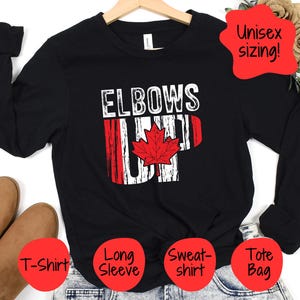 May include: Black long-sleeve t-shirt with the text "Elbows Up" and a red maple leaf graphic. The shirt is available in a variety of sizes and is unisex.