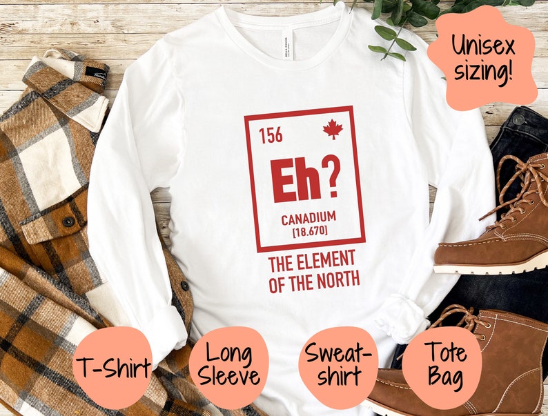 May include: White long-sleeve t-shirt with a red graphic of a periodic table element. The element is labeled "Eh?" with the text "Canadium [18.670]" and "The Element of the North". The shirt is shown on a wooden surface with a plaid shirt, a brown tote bag, and brown boots.