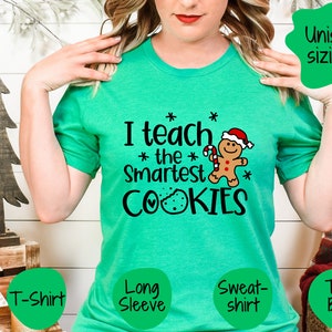 May include: Green t-shirt featuring the text "I teach the smartest cookies" and a gingerbread man graphic. The shirt is available in a variety of styles, including a t-shirt, long sleeve, sweatshirt, and tote bag.