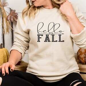 以下が含まれることがあります： 黒い文字で「hello fall」と書かれたベージュのトレーナー。fallの上には小さなハートと矢印があります。
