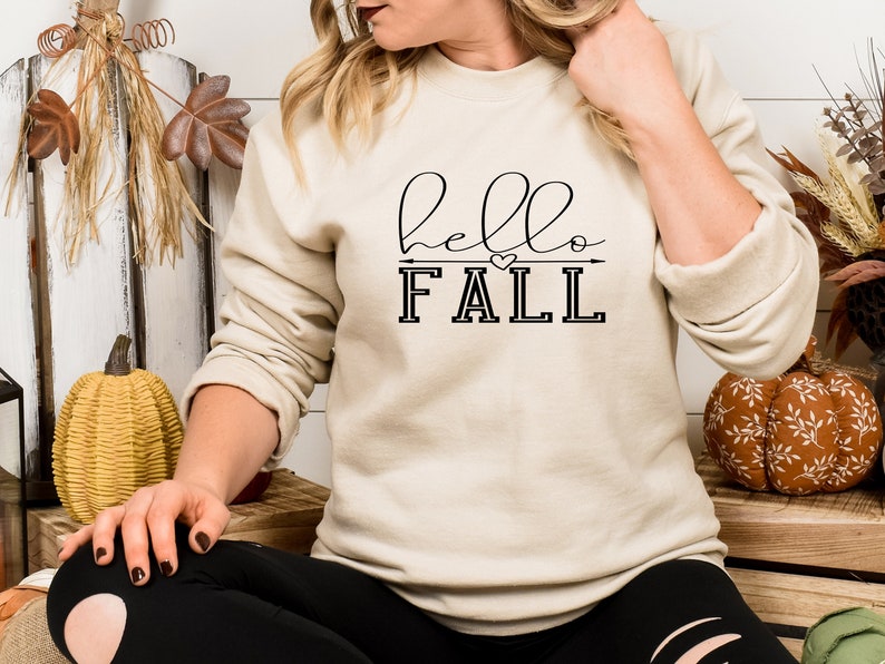以下が含まれることがあります： 黒い文字で「hello fall」と書かれたベージュのトレーナー。fallの上には小さなハートと矢印があります。
