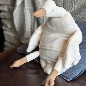 May include: A plush white duck toy with a beige beak and feet. The duck is sitting on a blue denim pillow. The toy has a soft, textured appearance, and its wings are outstretched. The background includes a patterned fabric and a brick wall.