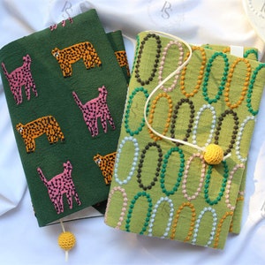 May include: Two green fabric book covers with different patterns. One cover features a repeating pattern of orange and black leopards and pink cats. The other cover has a repeating pattern of green, orange, and white circles.