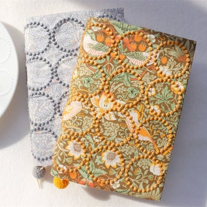 May include: Two fabric-covered journals with a floral pattern and a geometric pattern. The journals have a beaded trim and a tassel.