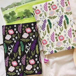 May include: Two embroidered book covers with floral patterns. One cover is black with pink and purple flowers, the other is white with pink and purple flowers. Both covers have green leaves and white accents.