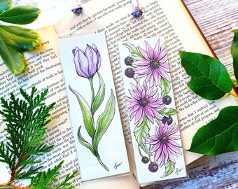 May include: Two handmade bookmarks featuring hand-painted floral designs. The first bookmark features a purple tulip with green leaves. The second bookmark features three pink flowers with purple centers and green leaves. Both bookmarks are on a white background.