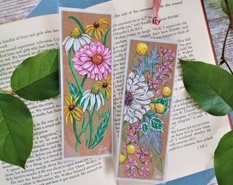 May include: Two brown paper bookmarks with floral illustrations. The first bookmark features pink, white, and yellow flowers on a brown background. The second bookmark features white, pink, and yellow flowers on a brown background with gray leaves. Both bookmarks have the word "Gin" written in black ink at the bottom.