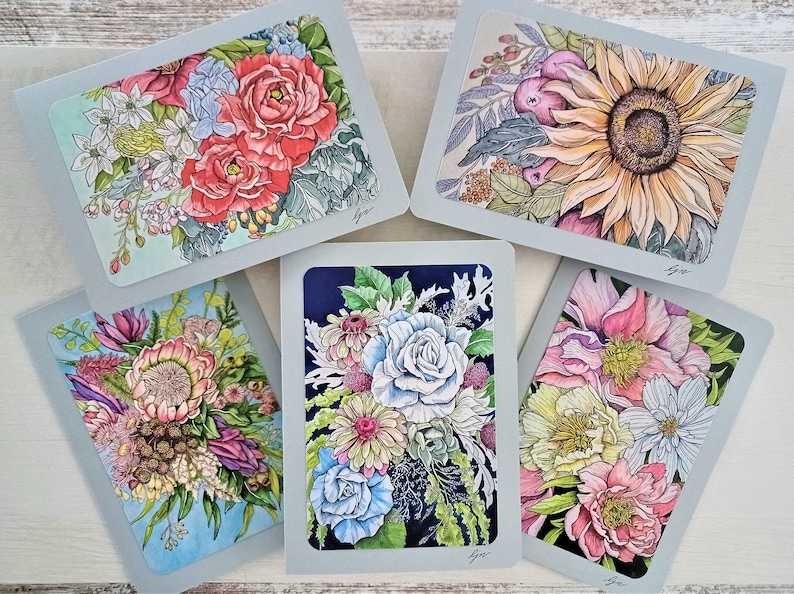 May include: Five greeting cards with floral illustrations. Each card features a different bouquet of flowers in various colors, including pink, blue, yellow, and white. The cards are all on a light wood background.