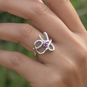Può includere: Anello color argento con un design astratto unico. L'anello presenta una piccola gemma rotonda color magenta al centro. L'anello è indossato su un dito, con uno sfondo verde sfocato.