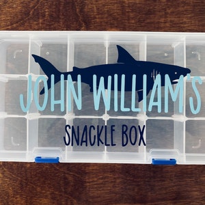 May include: A clear plastic storage box with multiple compartments. The box has a blue shark graphic and the text "JOHN WILLIAM'S SNACKLE BOX" printed on the front.