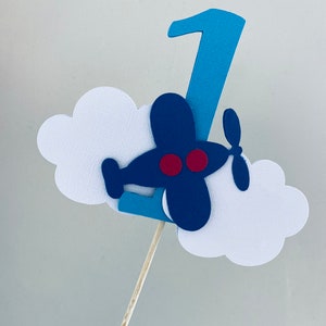 May include: Blue and white airplane cake topper with a red propeller and the number 1 on top. The airplane is flying over a white cloud.