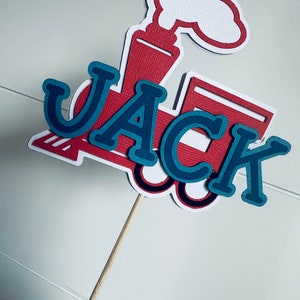 May include: A red, white, and blue train-shaped cake topper with the name "JACK" in layered blue letters. The topper is attached to a wooden stick for easy insertion into a cake or other dessert.