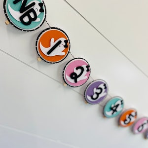 May include: A Halloween banner with a string of colourful circles featuring ghosts with black bows and numbers 1 through 6. The first circle is teal with the letters "NB" in black.