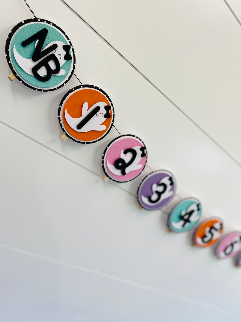 May include: A Halloween banner with a string of colourful circles featuring ghosts with black bows and numbers 1 through 6. The first circle is teal with the letters "NB" in black.