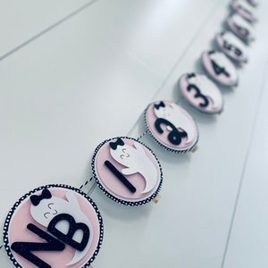 K&ouml;nnte beinhalten: Ein rosa-schwarzes Halloween-Banner mit wei&szlig;en Geistern und schwarzen Zahlen. Das Banner ist mit einer Schnur zusammengebunden und hat ein schwarz-wei&szlig;es Punkte-Muster.