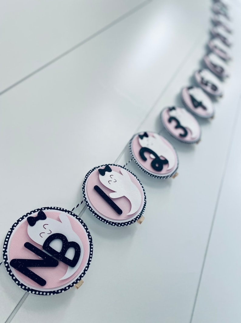 K&ouml;nnte beinhalten: Ein rosa-schwarzes Halloween-Banner mit wei&szlig;en Geistern und schwarzen Zahlen. Das Banner ist mit einer Schnur zusammengebunden und hat ein schwarz-wei&szlig;es Punkte-Muster.
