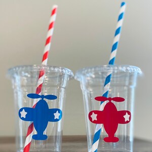 May include: Two clear plastic cups with red and blue striped straws. Each cup has a paper airplane sticker with a white star on each wing. One airplane is blue and the other is red.