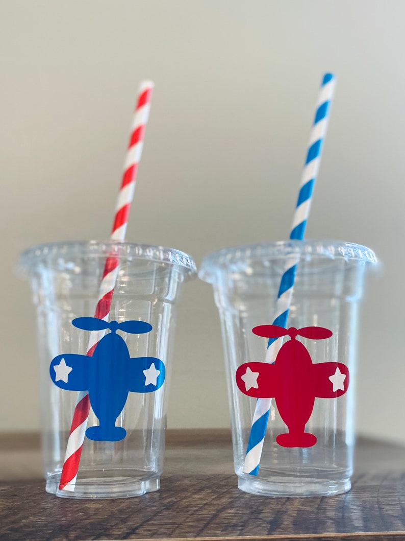 May include: Two clear plastic cups with red and blue striped straws. Each cup has a paper airplane sticker with a white star on each wing. One airplane is blue and the other is red.