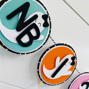 May include: A Halloween banner featuring three round paper circles. Each circle displays a white ghost with a black bow and a black letter on a coloured background. The first circle has a teal background with the letter "N", the second circle has an orange background with the letter "B", and the third circle has a pink background with the letter "1".
